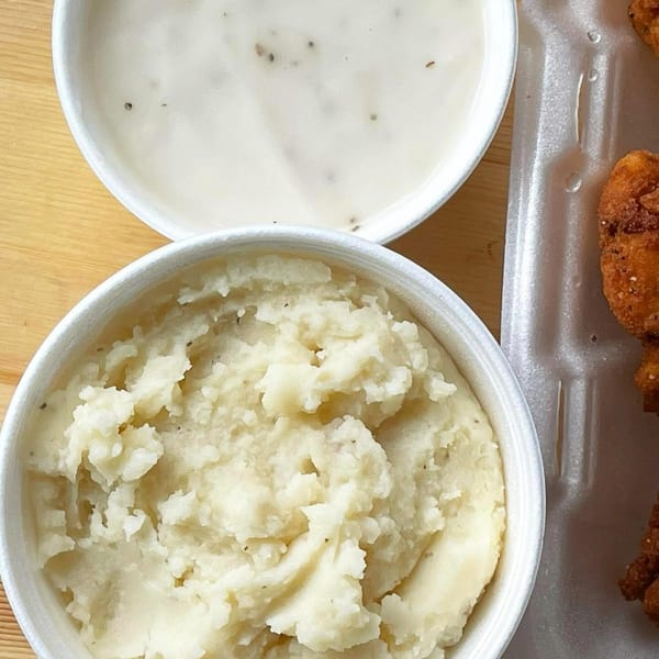 Creamy mashed potatoes and gravy in white bowls next to fried chicken in a takeout container.