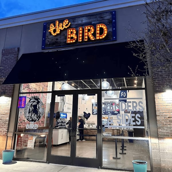 The Bird fried chicken restaurant exterior at dusk, with a lit-up sign and visible interior.