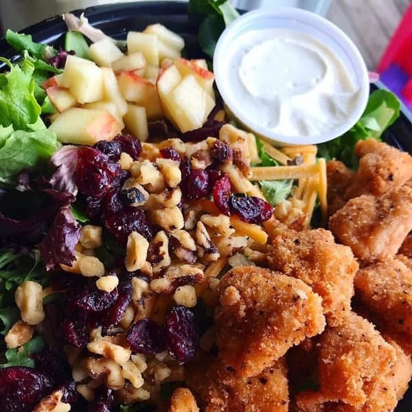 A fried chicken salad with mixed greens, apples, cranberries, walnuts, and a side of dressing.