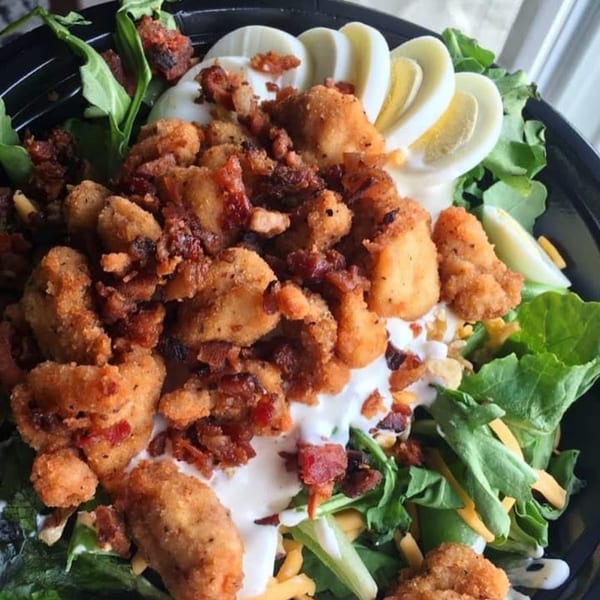 Fried chicken salad with bacon, hard-boiled eggs, cheese, and dressing in a black bowl.