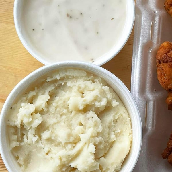 Creamy mashed potatoes and gravy in white bowls, next to fried chicken in a styrofoam container.